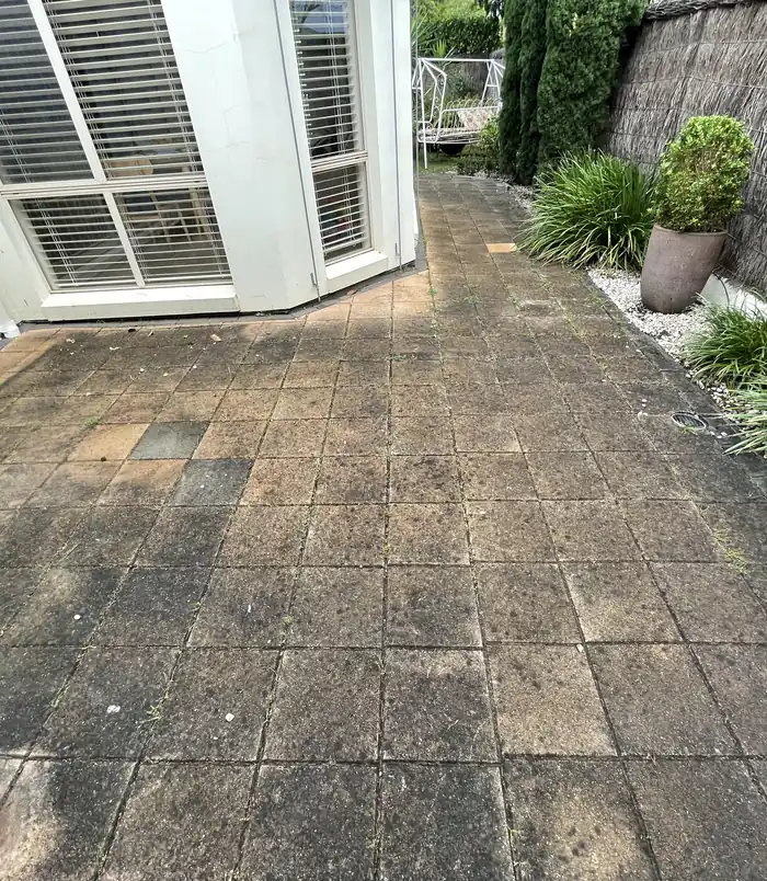 Dull, dirty pavers beside a white-framed bay window and garden, before pressure washing