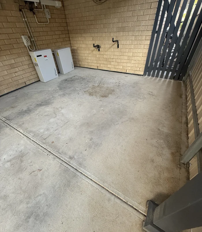 Pale, stained concrete courtyard beside a brick wall with water tanks, before resurfacing