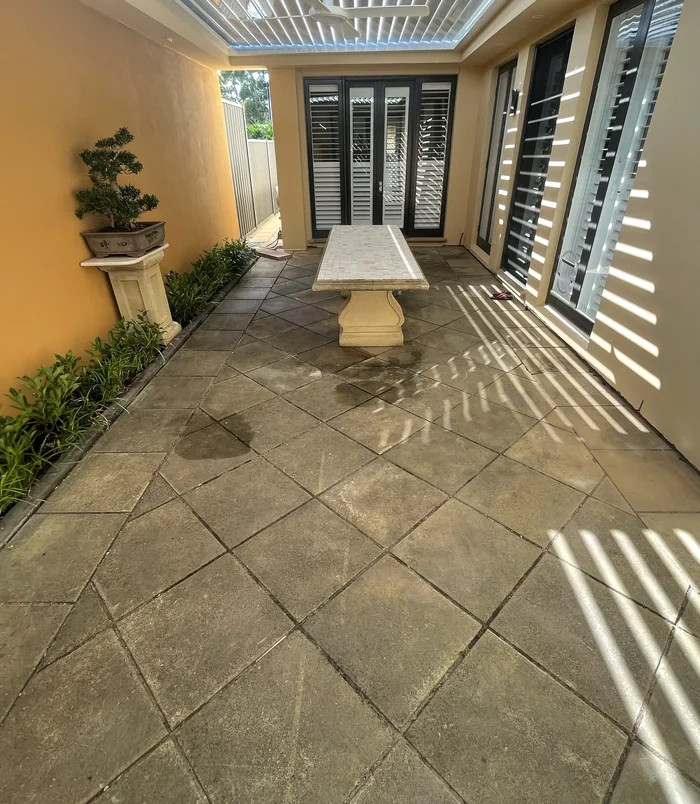 Dark, stained pavers in a covered courtyard with a stone table and shuttered doors, before pressure washing
