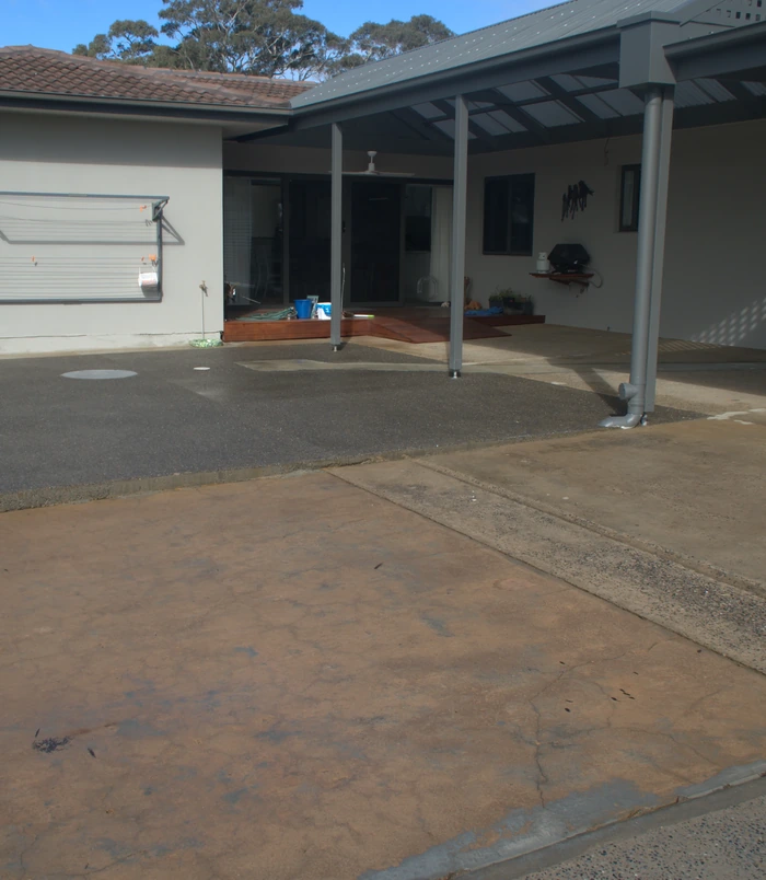 Stained, tired concrete patio under a pergola beside a house wall, before concrete resurfacing