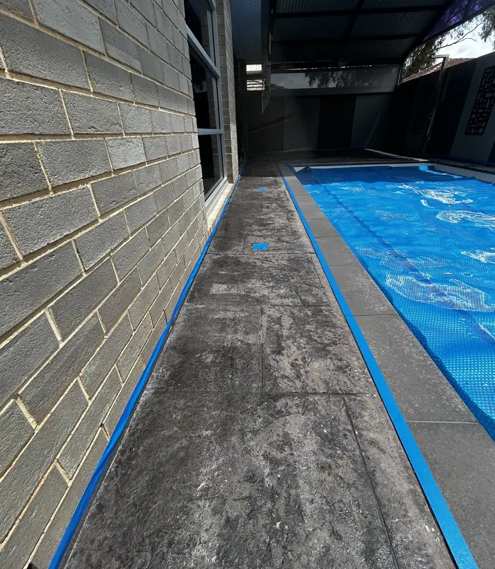 Stained concrete pool surround with blue masking tape beside a brick wall, before resurfacing