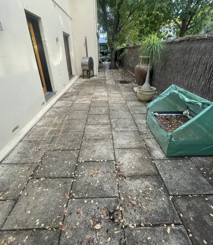 Dark, stained pavers in a side courtyard beside a rendered wall and garden bed, before pressure washing