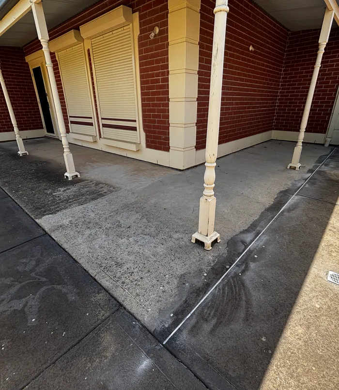 Worn concrete patio under a veranda beside a brick wall and roller shutters, before resurfacing