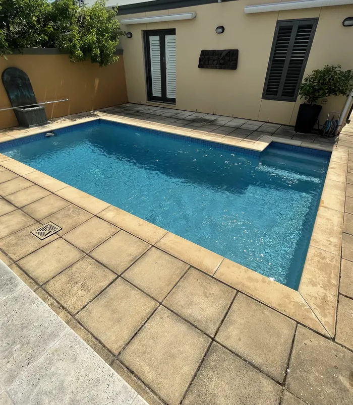 Dull, dirty pavers around a small blue swimming pool in a courtyard, before pressure washing