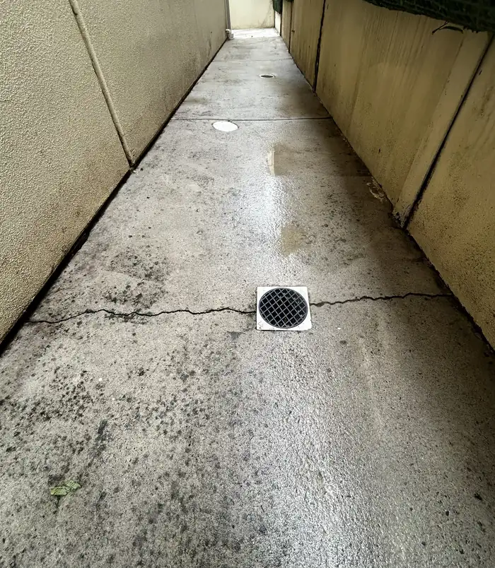 Cracked, stained concrete side path with a drain between two walls, before concrete resurfacing