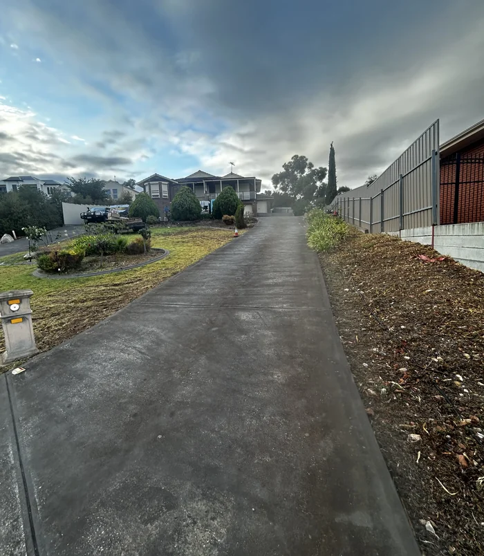 Worn, dark concrete driveway beside a tall fence under a cloudy sky, before concrete resurfacing