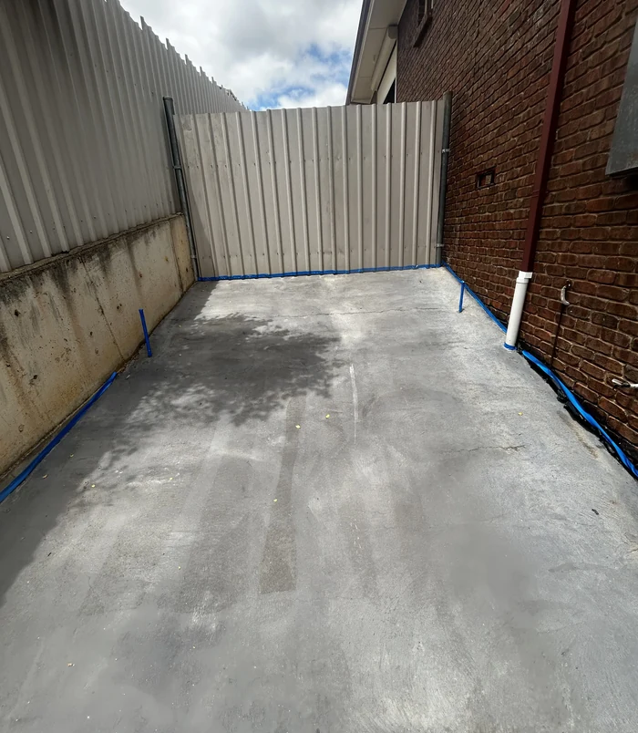Stained concrete courtyard with blue masking tape beside a brick wall and metal fence, before resurfacing