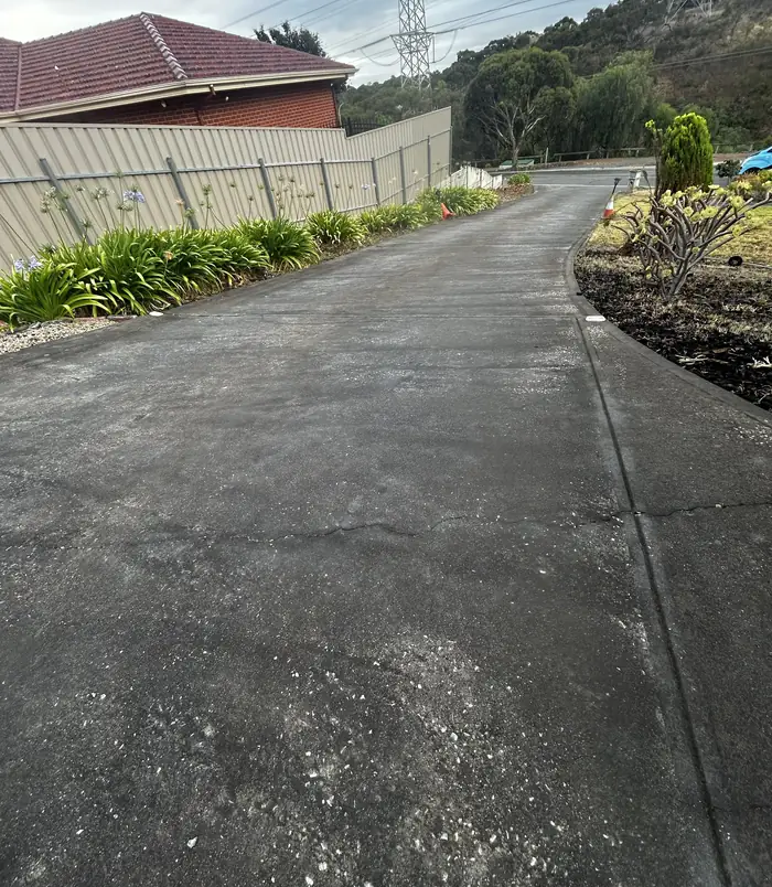 Dark, weathered concrete driveway curving beside a fence and garden, before concrete resurfacing