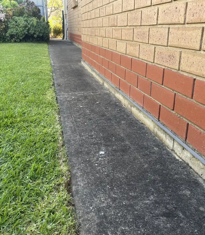 Dark, dirty concrete path beside a brick wall and lawn, before pressure washing