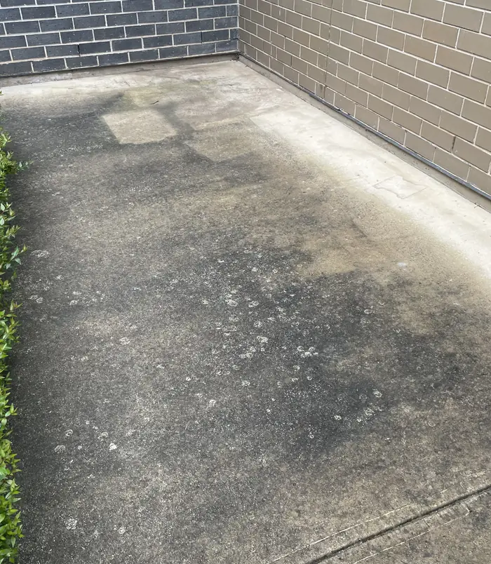 Stained, patchy concrete slab beside a grey brick wall, before pressure washing
