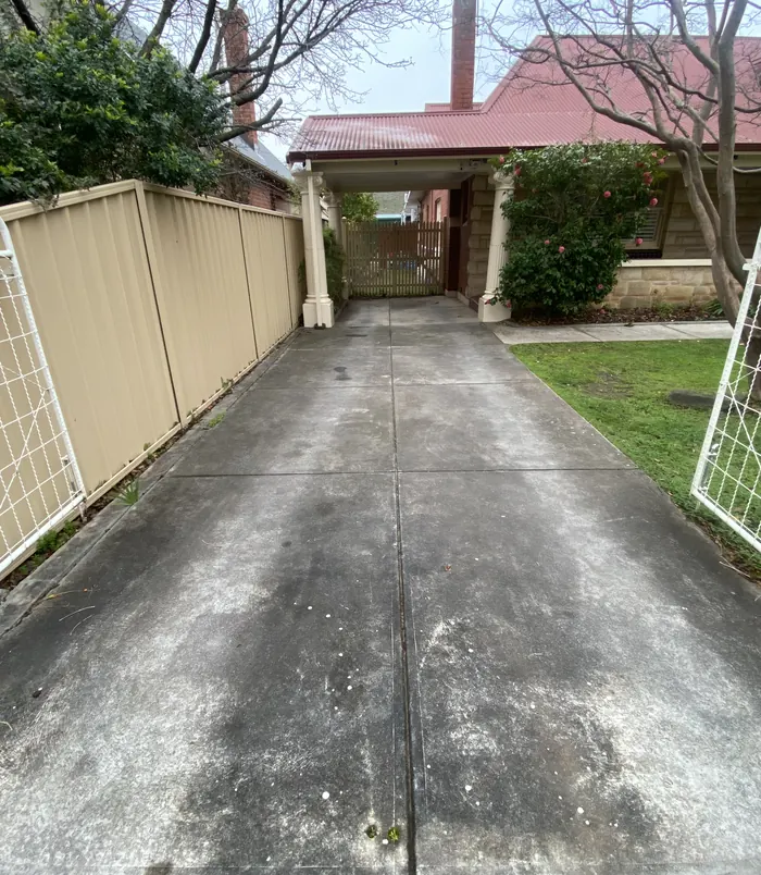 Stained, discoloured concrete driveway beside a tan fence and carport, before pressure washing
