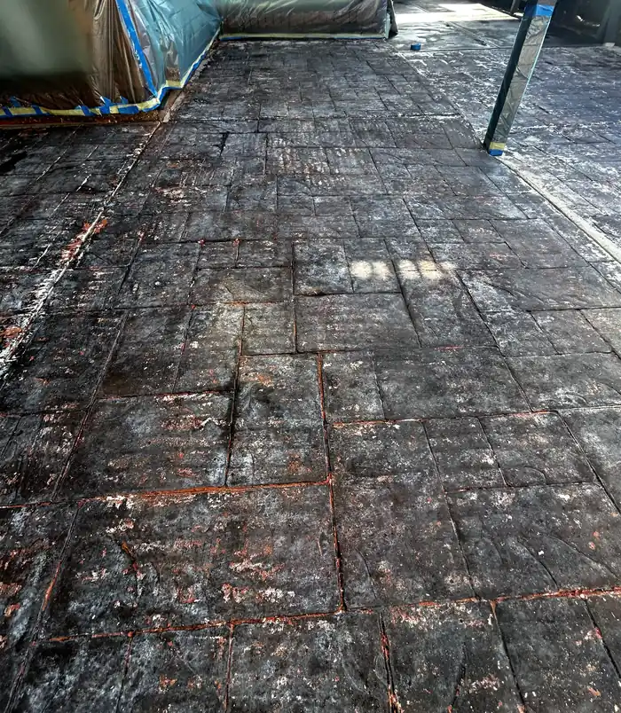 Stained, worn stamped-pattern concrete patio with blue tape along the edge, before resurfacing