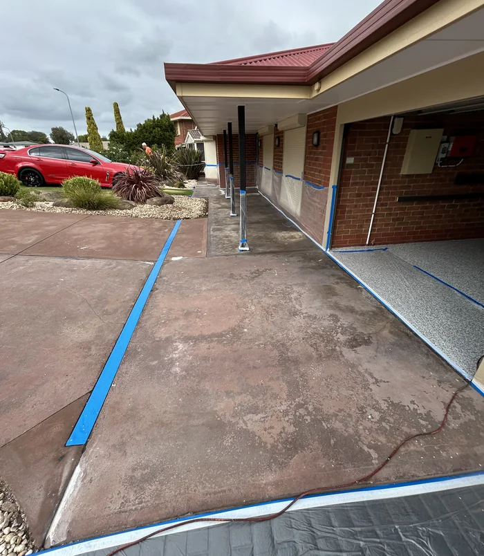 Faded, patchy red concrete path with blue masking tape beside a brick home with a red roof, before resurfacing
