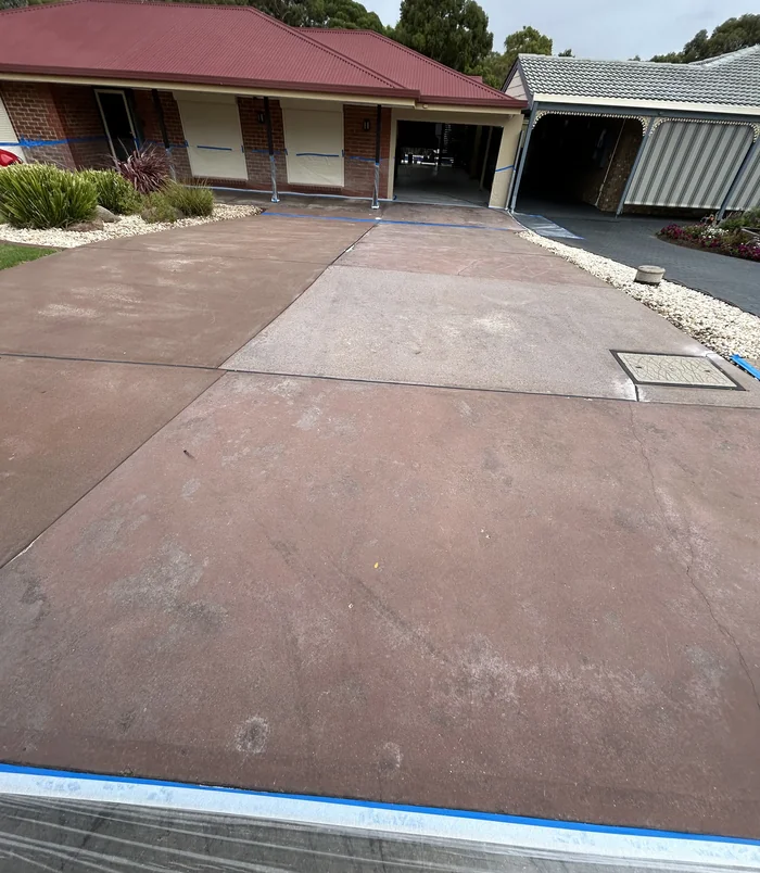 Faded, patchy pink concrete driveway with a drain cover in front of a brick home, before resurfacing