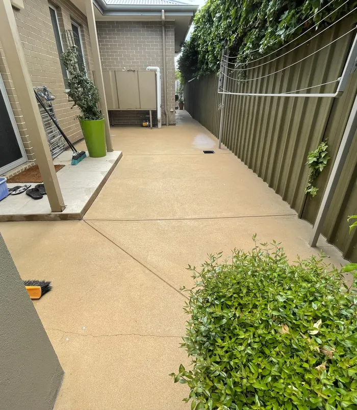 Clean, warm-toned concrete side path between a house and a green fence after pressure washing
