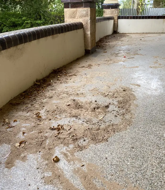 Dirty concrete walkway with mud and debris beside a rendered wall with sandstone pillars, before pressure washing