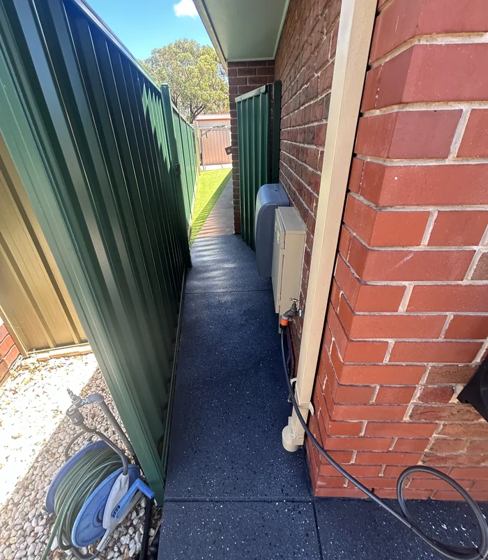Narrow side path beside a red brick wall and green fence with a smooth, dark resurfaced finish