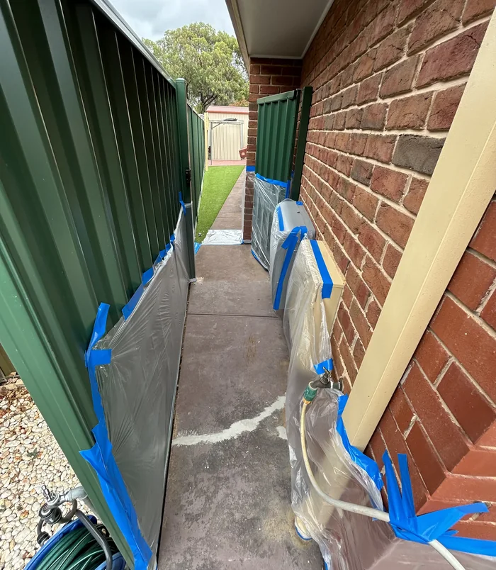 Narrow side path beside a brick wall and green fence with plastic and blue tape, before resurfacing
