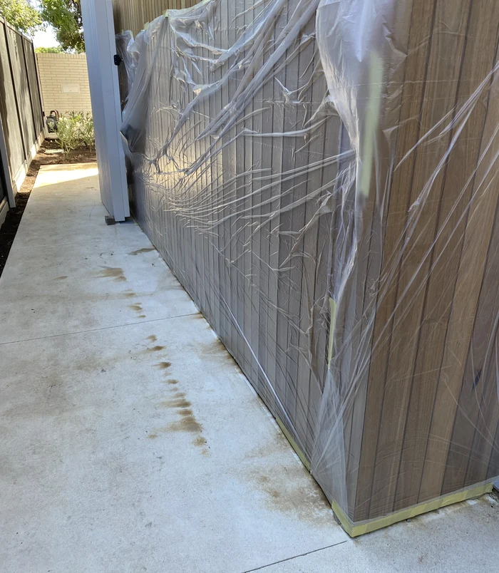 Dirty concrete side path beside a timber fence and a wall covered in plastic sheeting, before pressure washing