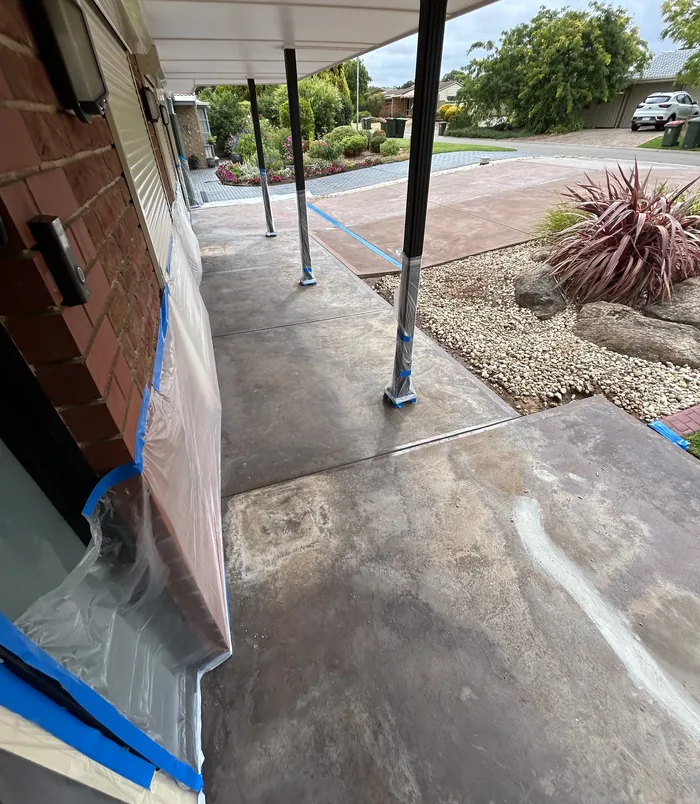 Stained concrete path with blue masking tape beside a brick wall and gravel garden, before resurfacing