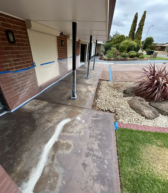 Stained concrete path with blue masking tape beside a brick wall and gravel garden, before resurfacing