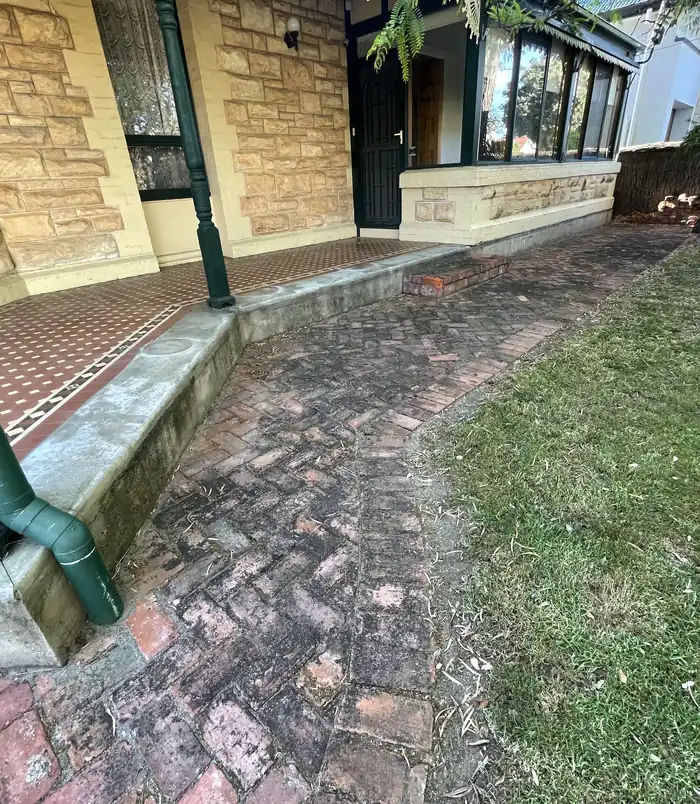 Dirty, algae-stained red brick paving beside a sandstone house, before pressure washing