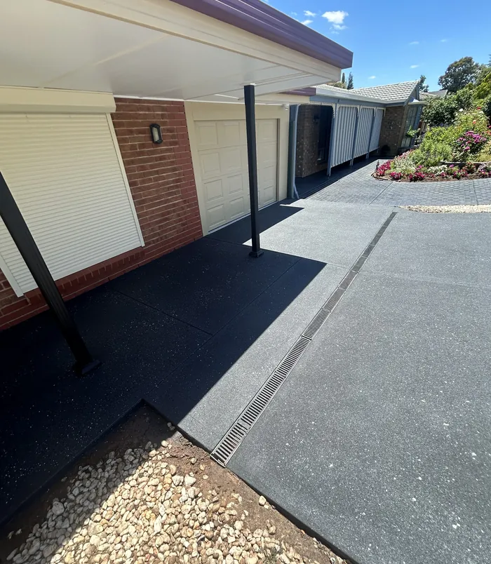 Dark grey resurfaced concrete path under a carport beside a brick wall