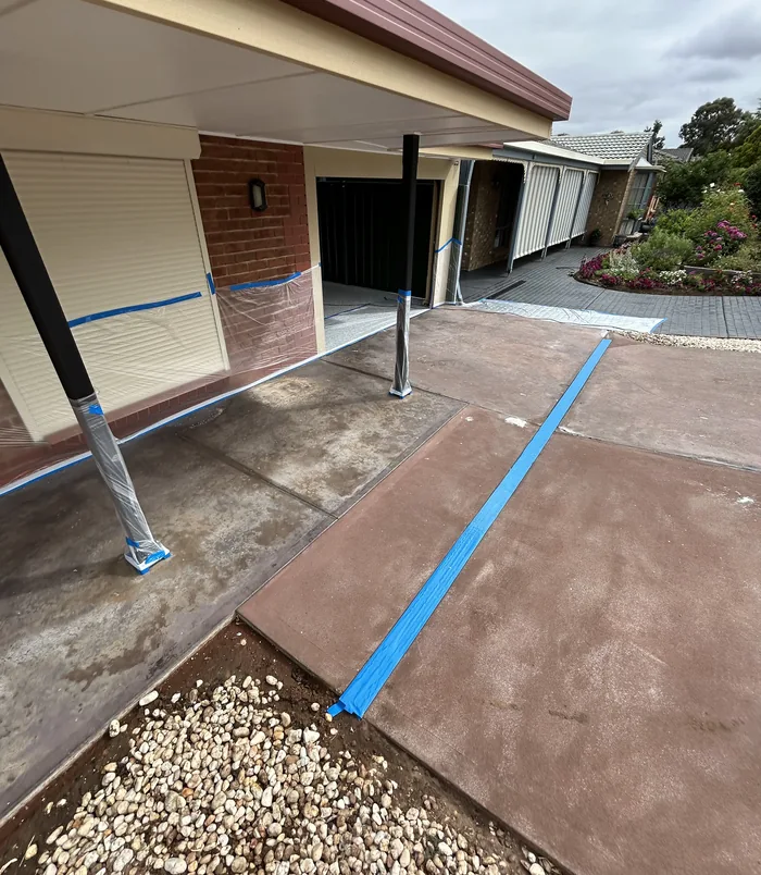 Faded red concrete path under a carport with blue masking tape beside a brick wall, before resurfacing
