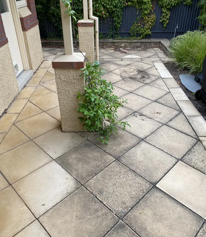 Stained, dirty diamond-pattern pavers around a stone pillar with a climbing plant, before pressure washing