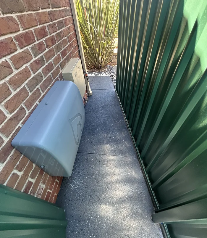 Narrow side path between a brick wall and green fence resurfaced in dark grey concrete