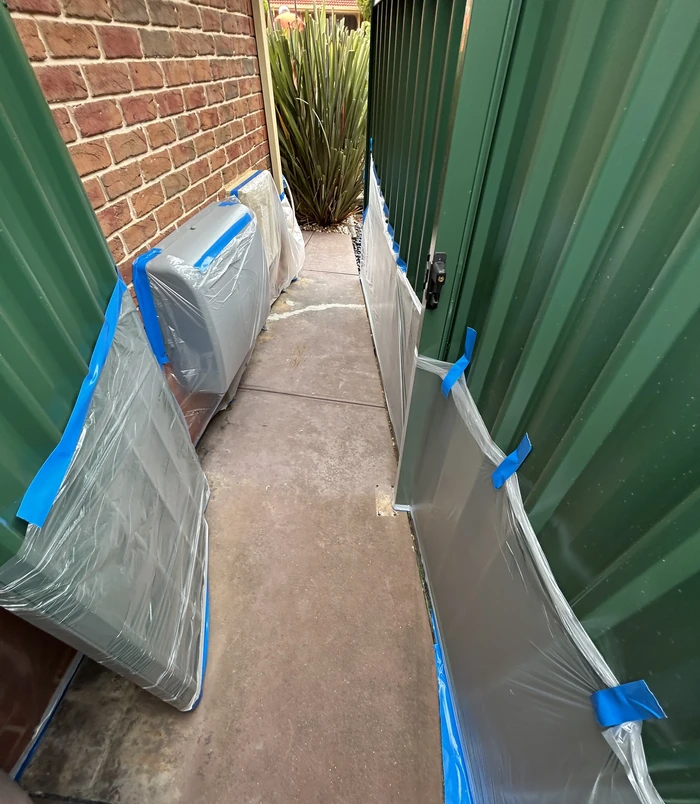 Narrow side path between a brick wall and green fence covered in plastic and blue tape, before resurfacing