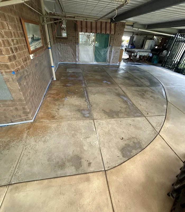 Patchy, stained concrete patio with blue masking tape beside a brick wall, before resurfacing