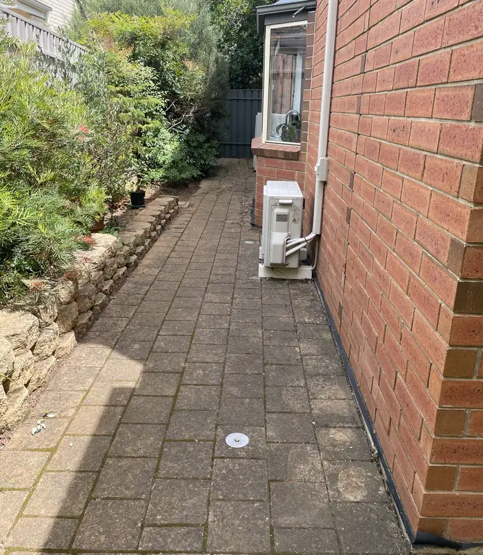 Dull, dirty brick pavers beside a red brick wall and air conditioner, before pressure washing