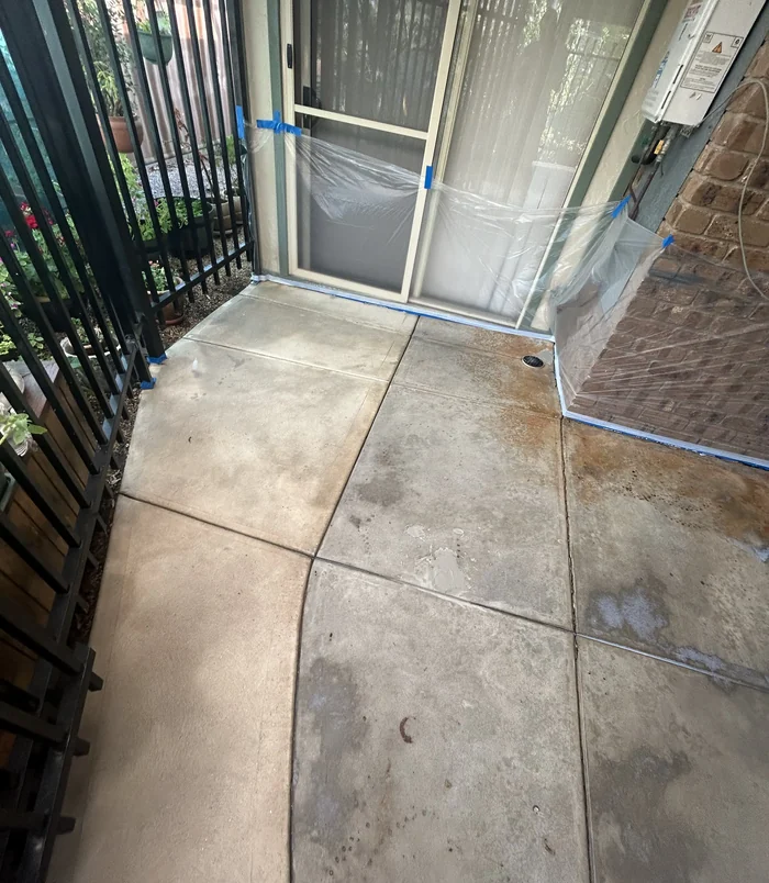 Stained, uneven concrete patio under a pergola beside a black fence, before concrete resurfacing
