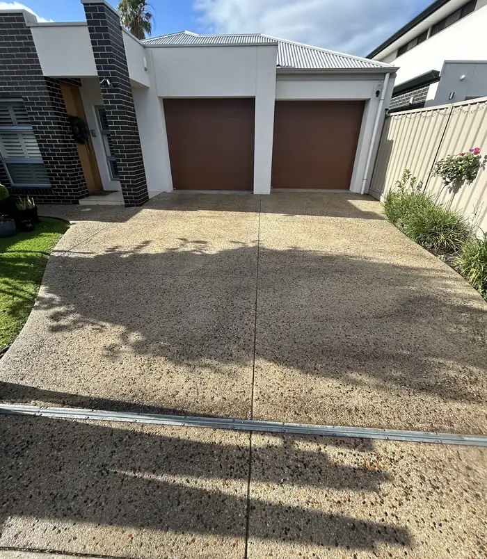 Pale, stained concrete driveway in front of a modern home with brown garage doors, before resurfacing