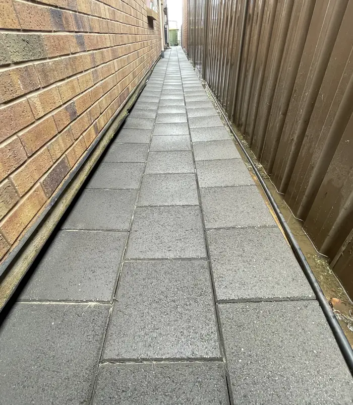 Narrow side passage between a brick wall and fence with clean, weed-free pavers after pressure washing