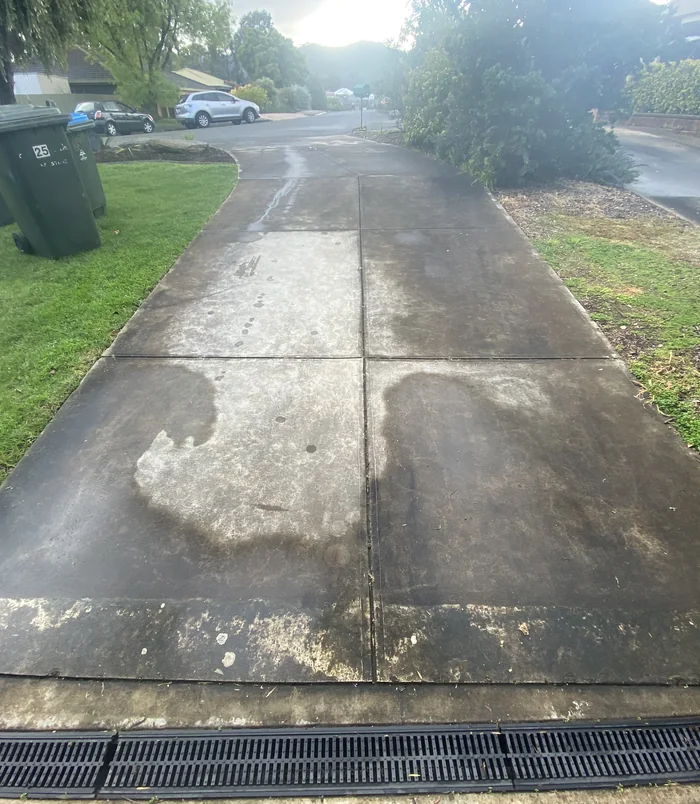 Concrete driveway with dark stains and grime beside a grass verge, before pressure washing
