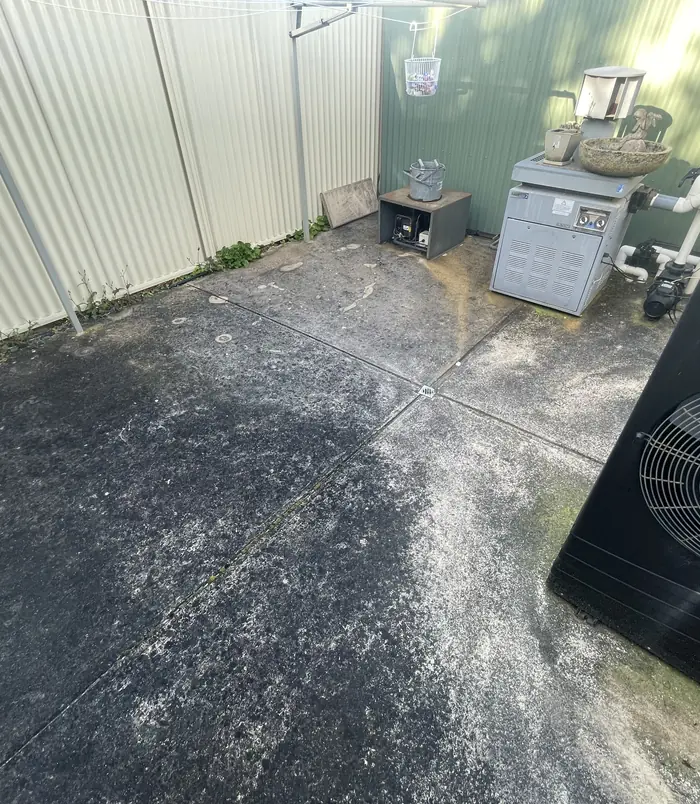 Dirty, stained concrete courtyard beside a fence with a pool pump and equipment, before pressure washing