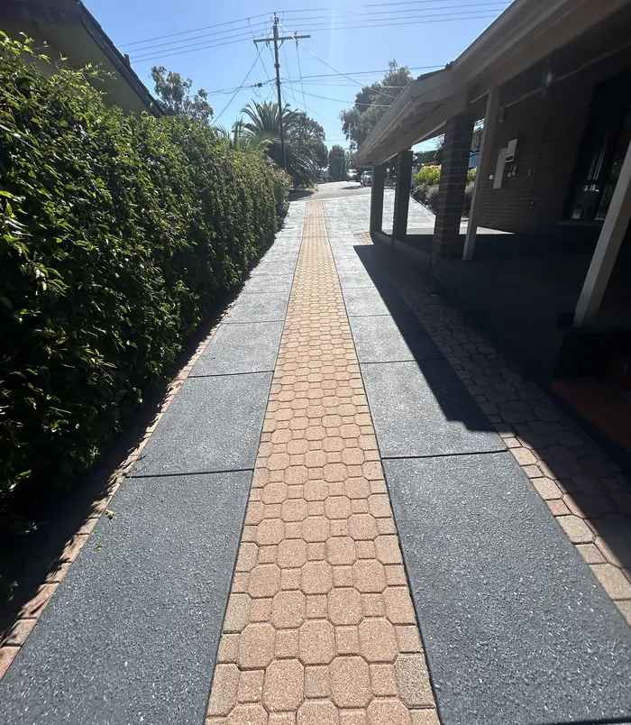 Dark grey resurfaced concrete path with a brick paver strip between a hedge and carport