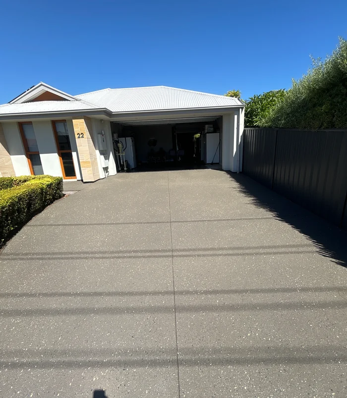 Dark grey resurfaced concrete driveway leading to a carport beside a hedge and fence