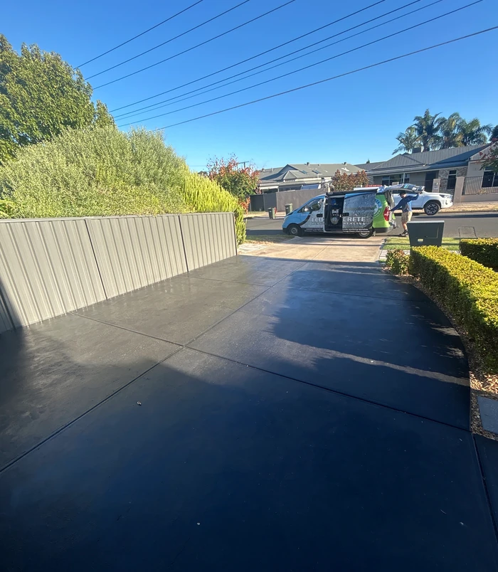 Dark, damp concrete driveway beside a metal fence and hedge, before concrete resurfacing