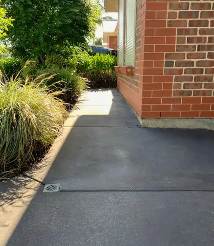 Worn grey concrete path beside a red brick wall and garden grasses, before concrete resurfacing