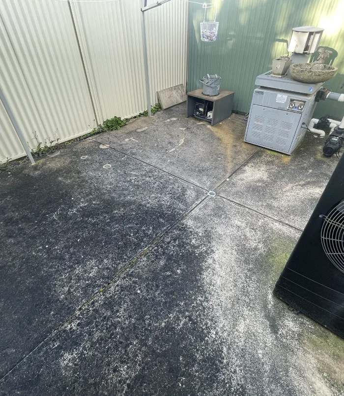 Dirty, stained concrete courtyard beside a fence with a pool pump and equipment, before concrete cleaning