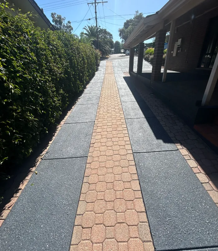 Dark grey resurfaced concrete path with a brick paver strip between a hedge and carport