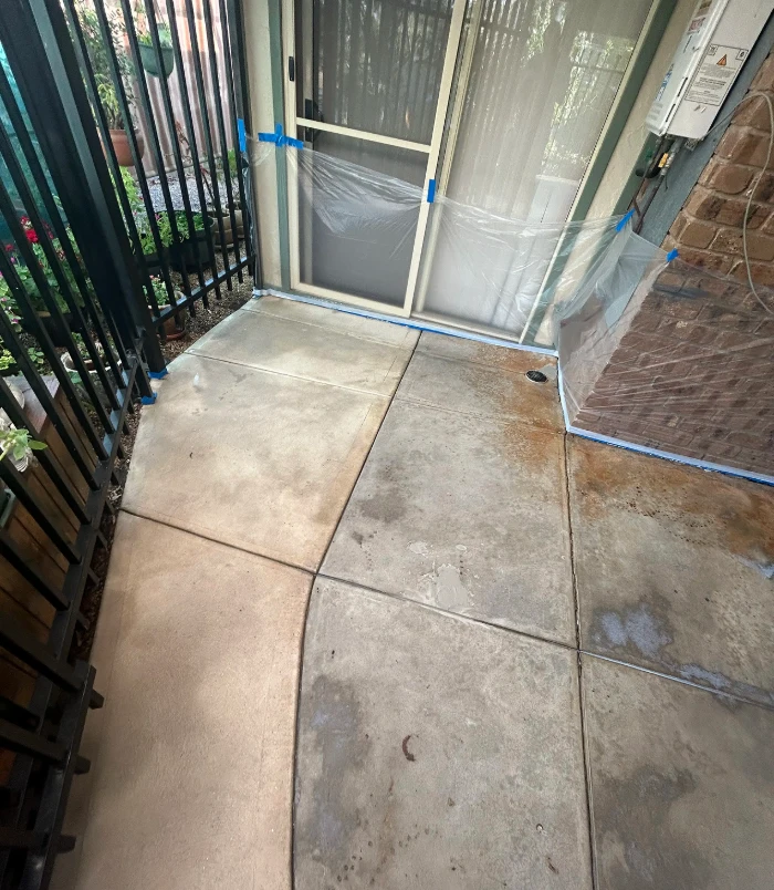 Stained concrete entry with blue masking tape beside a black fence and glass door, before resurfacing