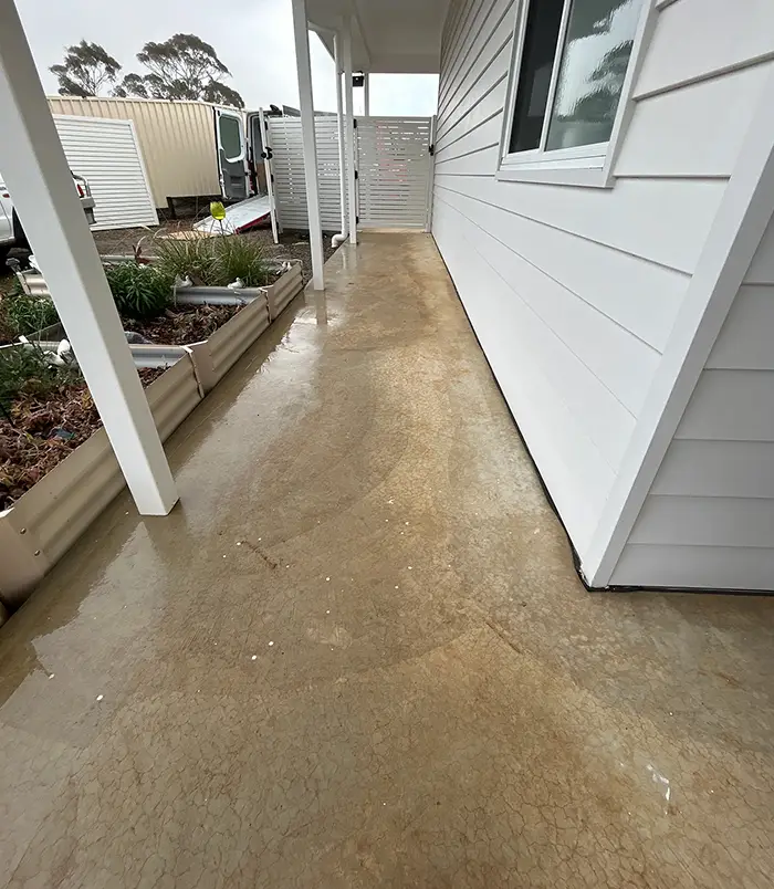 Stained, golden-brown concrete veranda floor beside a white weatherboard wall, before epoxy flooring