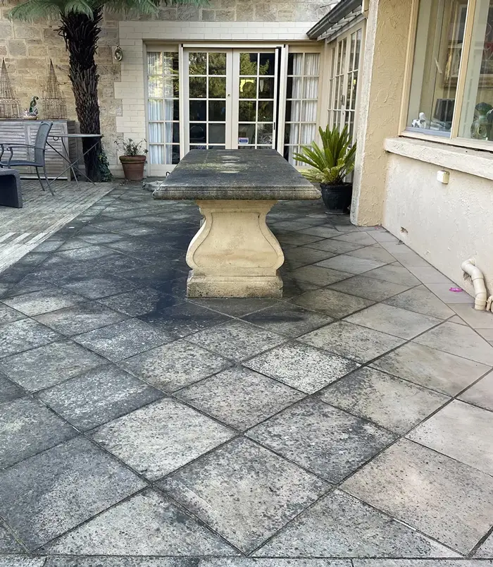 Dark, stained pavers around a stone outdoor table beside French doors, before paver cleaning