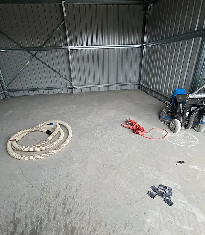 Bare concrete floor in a large metal shed with grinding equipment and a hose, before epoxy flooring