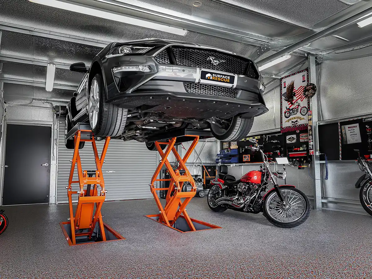 Dark sports car raised on orange hoists above a clean garage floor, with a motorcycle beside it