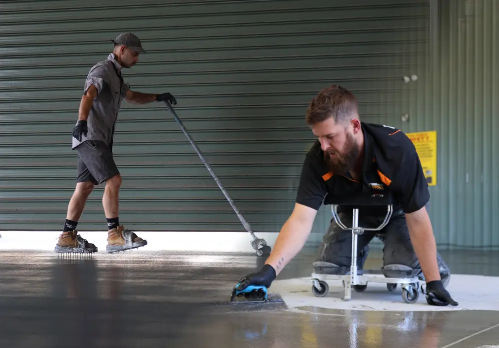 Two Surface Rescue technicians applying floor coating with long-handled tools in front of a roller door